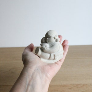 Op de afbeelding: Een kleine, cr&egrave;mekleurige keramische beeldje van een zittende figuur die een kleinere figuur vasthoudt. De figuren hebben afgeronde kenmerken en zitten. Het beeldje wordt in de hand van een persoon gehouden tegen een licht houten oppervlak.