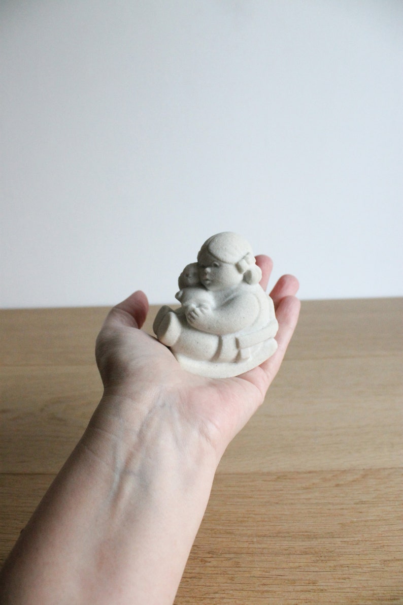 Op de afbeelding: Een kleine, cr&egrave;mekleurige keramische beeldje van een zittende figuur die een kleinere figuur vasthoudt. De figuren hebben afgeronde kenmerken en zitten. Het beeldje wordt in de hand van een persoon gehouden tegen een licht houten oppervlak.