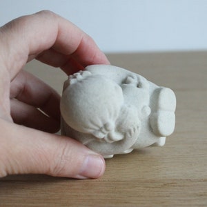 Op de afbeelding: Een kleine, gebroken witte keramische sculptuur van een varken met een afgerond lichaam en gedetailleerde kenmerken. Het varken wordt in een hand gehouden, waarbij het getextureerde oppervlak en het unieke ontwerp worden benadrukt. Decoratief item.
