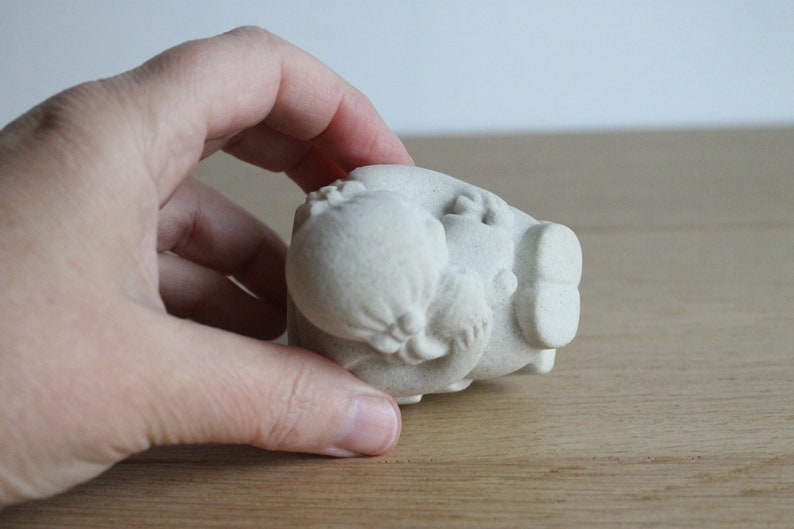 Op de afbeelding: Een kleine, gebroken witte keramische sculptuur van een varken met een afgerond lichaam en gedetailleerde kenmerken. Het varken wordt in een hand gehouden, waarbij het getextureerde oppervlak en het unieke ontwerp worden benadrukt. Decoratief item.