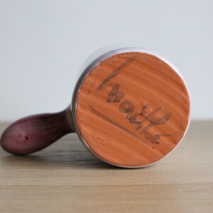 May include: The underside of a white ceramic mug with a brown handle. The base of the mug is terracotta and has the word "Martha" written on it.