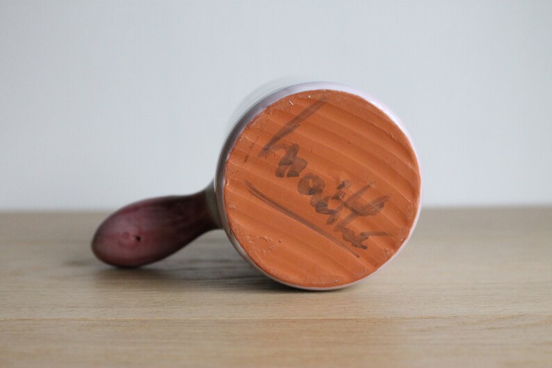 May include: The underside of a white ceramic mug with a brown handle. The base of the mug is terracotta and has the word "Martha" written on it.