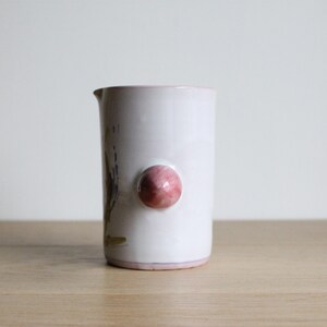 May include: White ceramic jug with a pink knob handle and a floral design. The jug has a slightly rounded shape and a wide mouth.