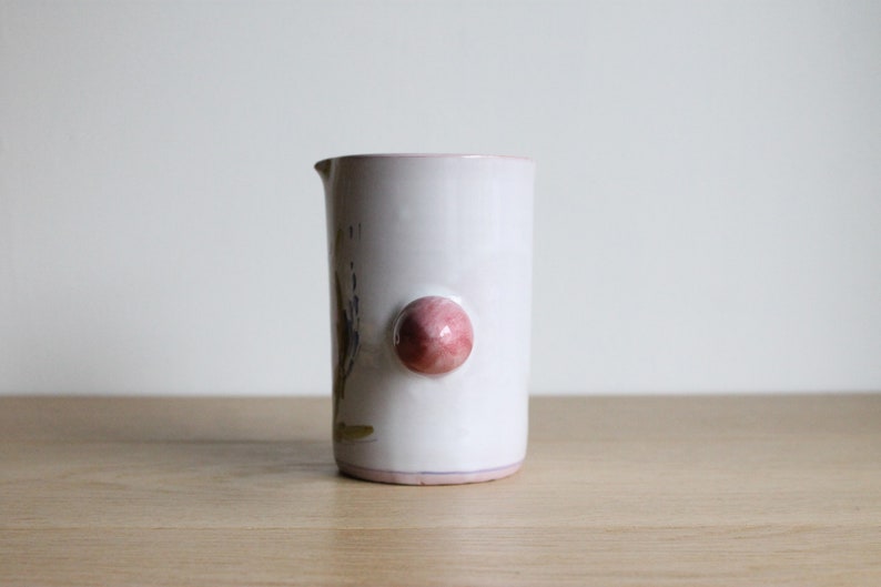 May include: White ceramic jug with a pink knob handle and a floral design. The jug has a slightly rounded shape and a wide mouth.