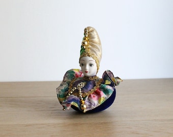 May include: A porcelain doll with a colorful, floral costume and a gold and green hat. The doll is sitting on a blue velvet base.