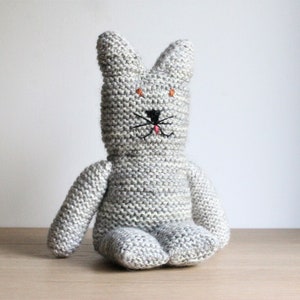 Könnte beinhalten: Ein graues, gestricktes Plüschtier in Form einer Katze mit schwarzen gestickten Augen und Schnurrhaaren. Das Spielzeug sitzt auf einer hellbraunen Holzoberfläche.