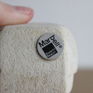 Op de afbeelding: Een lichtbeige stenen kunstsculptuur met een textuur. Een ronde sticker met de tekst "Marbell Stone Art Belgium" is op de sculptuur bevestigd. De sculptuur wordt door een vinger vastgehouden.