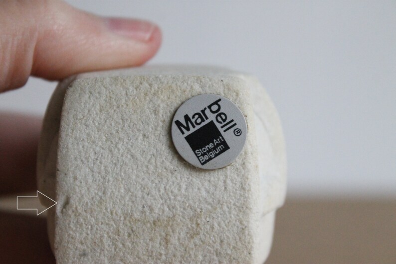 Op de afbeelding: Een lichtbeige stenen kunstsculptuur met een textuur. Een ronde sticker met de tekst "Marbell Stone Art Belgium" is op de sculptuur bevestigd. De sculptuur wordt door een vinger vastgehouden.