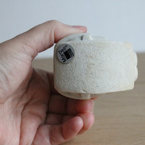 Op de afbeelding: Een lichtbeige, getextureerde, cilindrische sculptuur met een afgeronde boven- en onderkant. De sculptuur heeft een kleine, ronde sticker met de tekst "bell Marq". De sculptuur wordt in een hand gehouden.