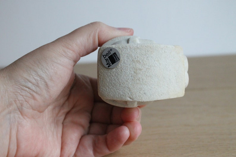 Op de afbeelding: Een lichtbeige, getextureerde, cilindrische sculptuur met een afgeronde boven- en onderkant. De sculptuur heeft een kleine, ronde sticker met de tekst "bell Marq". De sculptuur wordt in een hand gehouden.