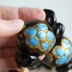 Puede incluir: Primer plano de un collar negro y dorado con cuentas pintadas de azul y dorado. Las cuentas son redondas y tienen un acabado brillante.
