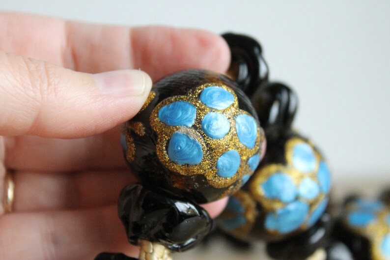 Puede incluir: Primer plano de un collar negro y dorado con cuentas pintadas de azul y dorado. Las cuentas son redondas y tienen un acabado brillante.