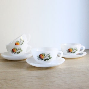 May include: White teacups and saucers with a fruit design. The cups are stacked and arranged on a light-colored wooden surface. Each cup and saucer features a colorful illustration of a peach and green leaves.
