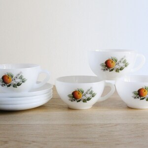 May include: A set of white ceramic teacups and saucers. Each cup and saucer features a painted design of an orange fruit with green leaves. The cups have handles and are arranged on a wooden surface.