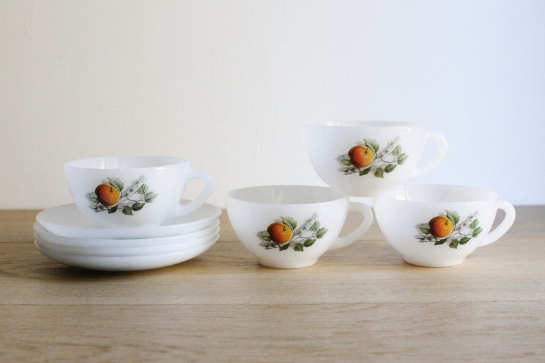May include: A set of white ceramic teacups and saucers. Each cup and saucer features a painted design of an orange fruit with green leaves. The cups have handles and are arranged on a wooden surface.