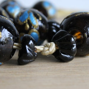 Puede incluir: Primer plano de un collar de cuentas negras y doradas con detalles azules. Las cuentas son de vidrio y tienen un acabado brillante. El collar est&aacute; enhebrado en un cord&oacute;n de color crema.