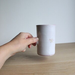 May include: A white ceramic mug with a pink flower design. The mug is being held by a hand.