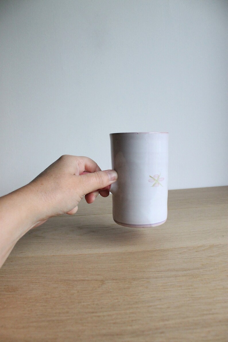 May include: A white ceramic mug with a pink flower design. The mug is being held by a hand.
