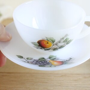 May include: White teacup and saucer set with a fruit design. The teacup and saucer are white with a fruit design featuring an orange peach, green leaves, and purple grapes. The teacup has a handle and sits on a matching saucer.