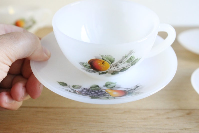 May include: White teacup and saucer set with a fruit design. The teacup and saucer are white with a fruit design featuring an orange peach, green leaves, and purple grapes. The teacup has a handle and sits on a matching saucer.