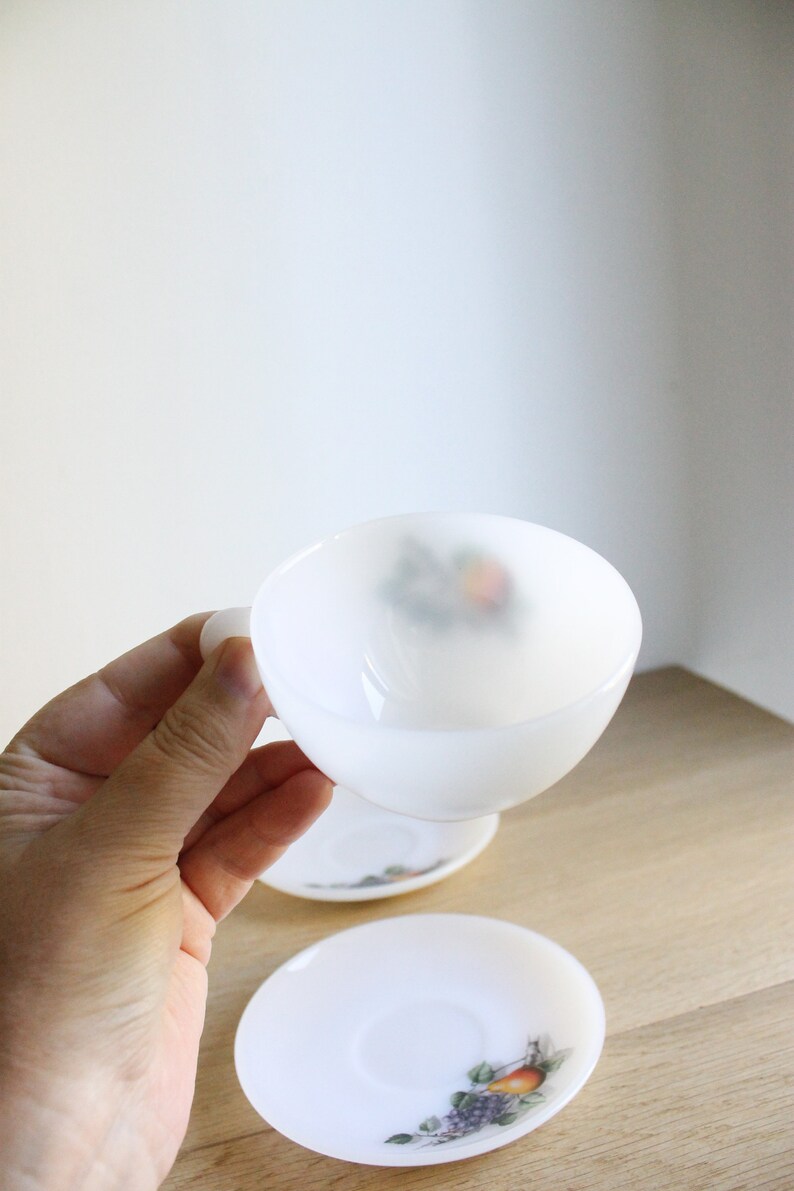 May include: White milk glass teacup and saucer set. The cup is held in a hand, with a matching saucer below. Both pieces feature a fruit design in the center. The set is on a wooden surface.