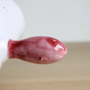 May include: A close-up of a red ceramic handle on a white ceramic dish. The handle is smooth and glossy, with a slight pink hue.