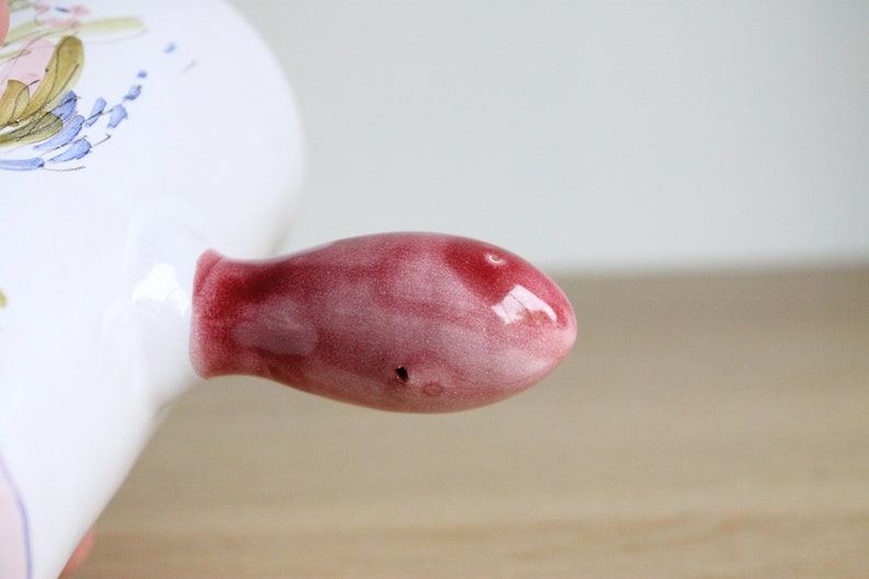May include: A close-up of a red ceramic handle on a white ceramic dish. The handle is smooth and glossy, with a slight pink hue.