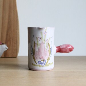 May include: A white ceramic jug with a pink floral design and a pink handle. The jug is on a wooden surface.