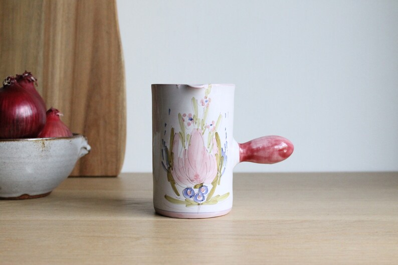 May include: A white ceramic jug with a pink floral design and a pink handle. The jug is on a wooden surface.
