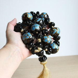Puede incluir: Un collar largo de cuentas negras y doradas con detalles azules y un borla dorada. Las cuentas son redondas y tienen un dise&ntilde;o floral.