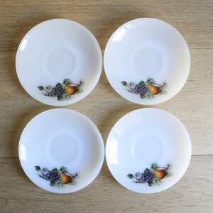 May include: Four white, round saucers with a fruit design. Each saucer features a central depression and a printed image of grapes, a pear, and green leaves. The saucers are arranged on a light-colored wooden surface.