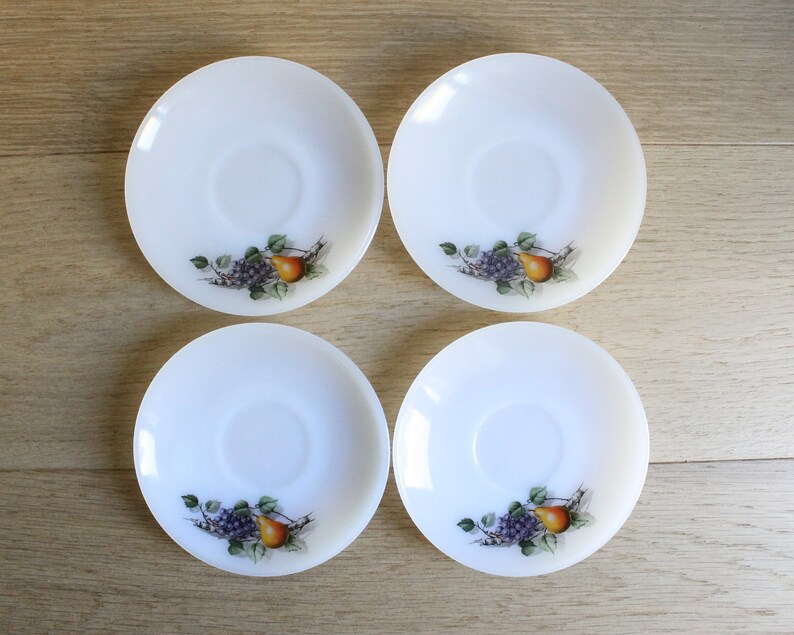 May include: Four white, round saucers with a fruit design. Each saucer features a central depression and a printed image of grapes, a pear, and green leaves. The saucers are arranged on a light-colored wooden surface.