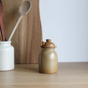 Può includere: Un barattolo di ceramica marrone con un coperchio di legno e un pomello di legno. Il barattolo è su una superficie di legno.