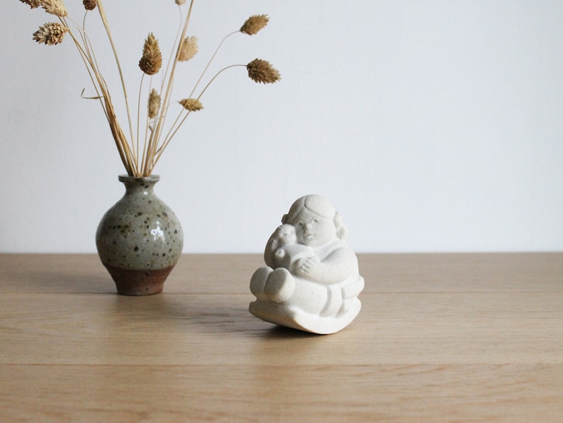 Op de afbeelding: Een kleine, cr&egrave;mekleurige keramische figuur van een kind dat een teddybeer vasthoudt, zittend op een schommelbasis. Links staat een kleine, gespikkelde groen-bruine vaas met gedroogde tarwestengels.