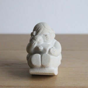 Op de afbeelding: Een kleine, gebroken witte beeldje van een zittende figuur die een kleinere figuur vasthoudt. De sculptuur heeft een matte afwerking en staat tegen een lichte achtergrond. De figuur draagt een jurk en heeft een strik in haar haar.
