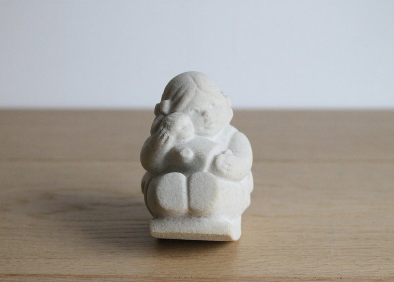 Op de afbeelding: Een kleine, gebroken witte beeldje van een zittende figuur die een kleinere figuur vasthoudt. De sculptuur heeft een matte afwerking en staat tegen een lichte achtergrond. De figuur draagt een jurk en heeft een strik in haar haar.