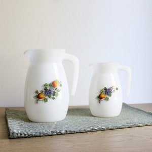 Könnte beinhalten: Zwei weiße Keramikkrüge mit einem Fruchtdesign. Der größere Krug hat eine Birne, Trauben und einen Apfel. Der kleinere Krug hat eine Birne und Trauben. Beide Krüge haben einen weißen Hintergrund mit einem grünen Rankendesign.