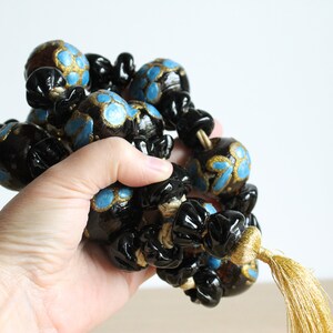 Puede incluir: Una guirnalda de cuentas negras y doradas con detalles azules y un borla dorada. Las cuentas son redondas y tienen un dise&ntilde;o floral.