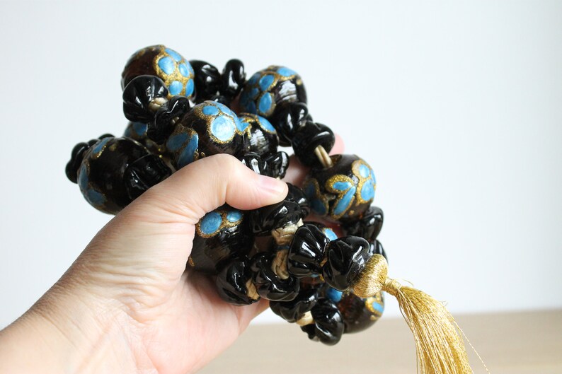 Puede incluir: Una guirnalda de cuentas negras y doradas con detalles azules y un borla dorada. Las cuentas son redondas y tienen un dise&ntilde;o floral.