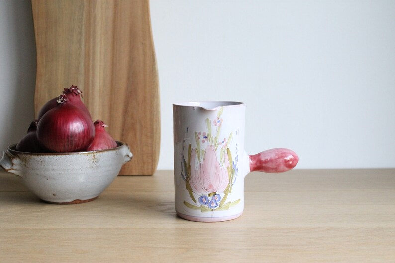 May include: A white ceramic jug with a pink handle and floral design. The jug features a pink flower with blue accents and green leaves.