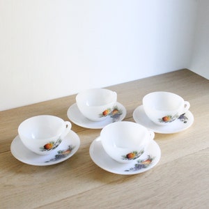 May include: Four white teacups and saucers are arranged on a light wood surface. Each saucer and cup features a colorful fruit and grape design. The cups have small handles.