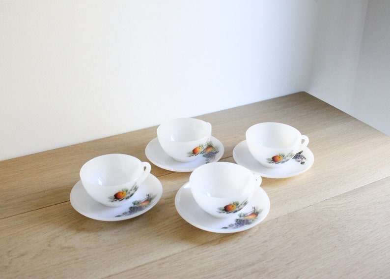 May include: Four white teacups and saucers are arranged on a light wood surface. Each saucer and cup features a colorful fruit and grape design. The cups have small handles.