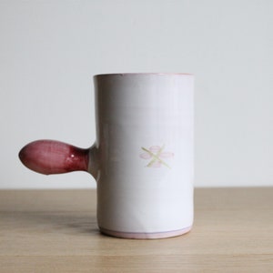 May include: White ceramic mug with a pink handle and a pink flower design. The mug has a light pink rim.