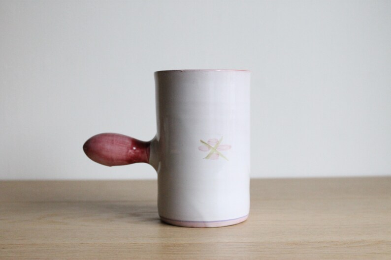 May include: White ceramic mug with a pink handle and a pink flower design. The mug has a light pink rim.