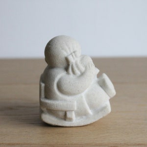 Op de afbeelding: Een kleine, gebroken witte figuur van een zittende figuur, mogelijk een monnik of godheid, met een serene uitdrukking. De figuur houdt een bundel vast en de details zijn subtiel gesneden. De sculptuur staat op een houten oppervlak.