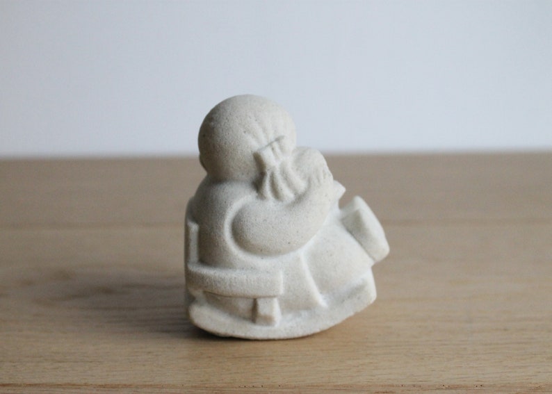 Op de afbeelding: Een kleine, gebroken witte figuur van een zittende figuur, mogelijk een monnik of godheid, met een serene uitdrukking. De figuur houdt een bundel vast en de details zijn subtiel gesneden. De sculptuur staat op een houten oppervlak.