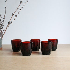 Può includere: Set di sei bicchieri da bere in vetro rosso scuro. I bicchieri hanno un design sfaccettato e sono disposti in fila su una superficie in legno.