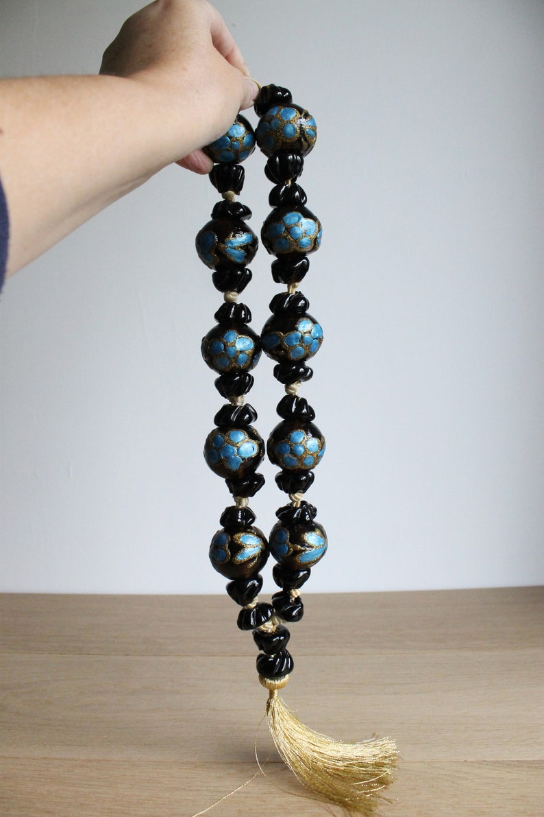 Puede incluir: Un collar de cuentas negras con detalles azules y dorados. El collar tiene un borla al final.