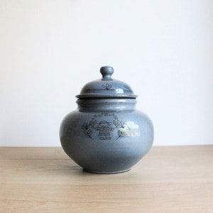 Peut inclure: Un pot en céramique bleu-gris avec un couvercle. Le pot a un corps rond et un petit bouton sur le dessus du couvercle. Un motif décoratif d'enfant et d'éléments floraux entoure le pot. Le pot est posé sur une surface en bois.