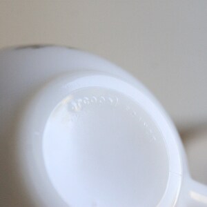 May include: Close-up of a white, opaque, ceramic mug. The bottom of the mug is visible, with the word "arcoos" embossed on the base. The mug has a rounded shape and a handle is partially visible.